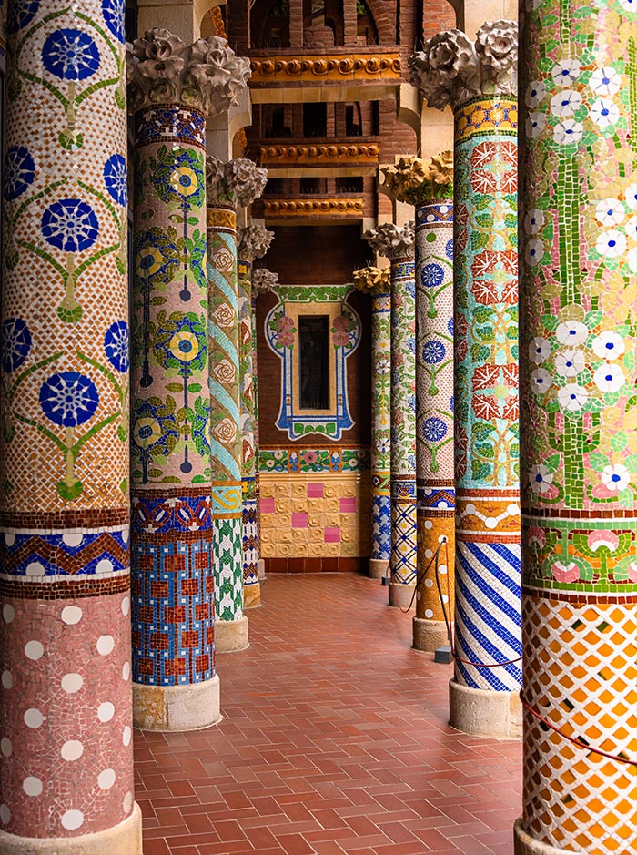 Colorful mosaic-covered columns on the balcony of the Palau de la Música Catalana in Barcelona, featuring intricate floral and geometric patterns in vibrant blues, greens, yellows, and reds. The red-tiled floor and decorative wall details add to the ornate, modernist design of the space.