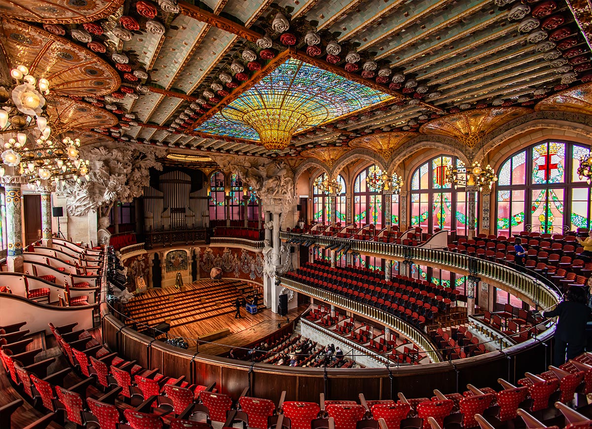 Ornate concert hall interior of the Palau de la Música Catalana in Barcelona, with a vibrant stained glass skylight, detailed sculptural columns, red velvet seating, and large stained glass windows lining the side walls.







