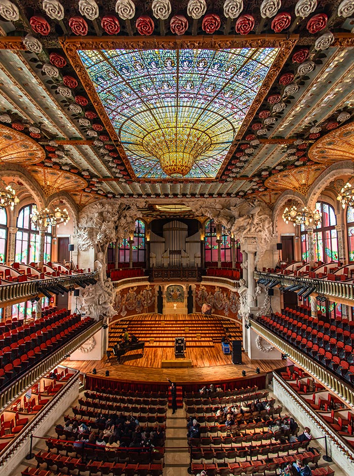 View of the Palau de la Música Catalana concert hall in Barcelona from the back of the auditorium, showcasing the colorful stained glass skylight, intricate ceiling decor with rose motifs, and the grand stage framed by sculptural columns and a pipe organ. Red and gold seating wraps around the hall, with light streaming in through stained glass windows.