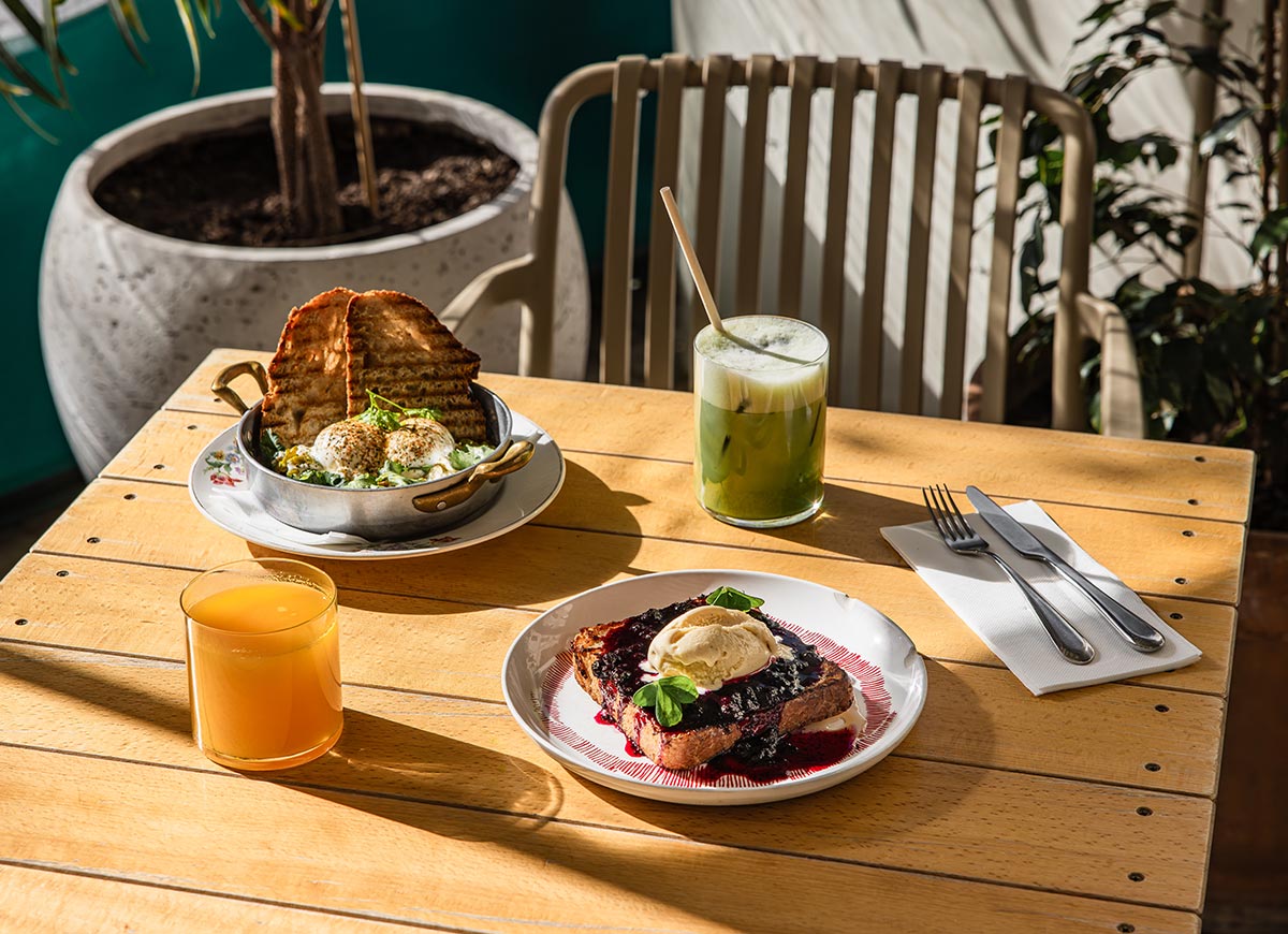 Sunny brunch setup at Salut Bistro in Barcelona, featuring a dish of poached eggs with toast, a plate of French toast topped with berry compote and ice cream, and two drinks—one green iced matcha and one orange juice—on a wooden table with natural light streaming in.