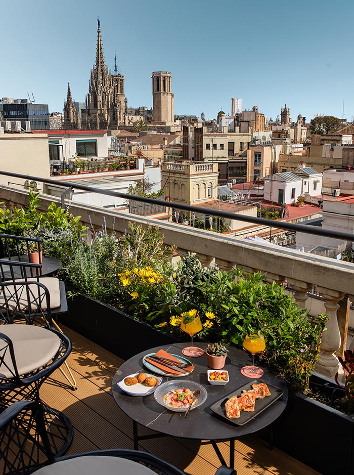 Charming rooftop terrace at Terraza de Vivi in Barcelona, set with a round table featuring tapas like pan con tomate, croquettes, ceviche, and fresh fruit, along with vibrant cocktails. The terrace overlooks a sweeping cityscape with the Gothic Cathedral of Barcelona rising prominently in the background. Surrounded by flowering plants and stylish black chairs, the scene exudes a relaxed, scenic dining vibe.