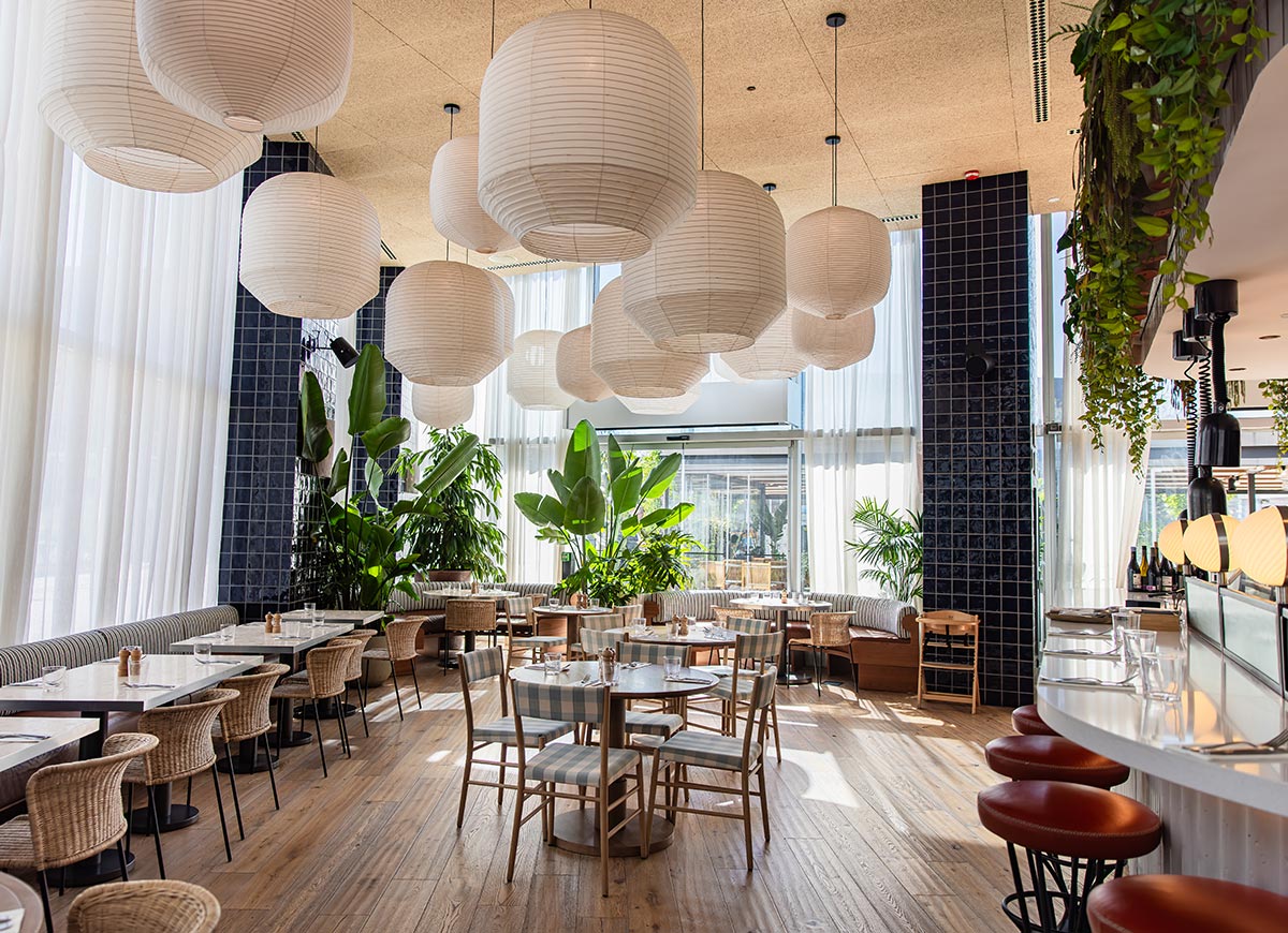 Airy and stylish restaurant interior with tall white curtains, hanging paper lanterns, and lush green plants throughout the space. The seating includes a mix of wicker and upholstered chairs around marble tables, with a curved bar area featuring red leather stools on the right. Natural light floods the room, highlighting the wood floors and navy tiled columns for a relaxed, modern vibe.