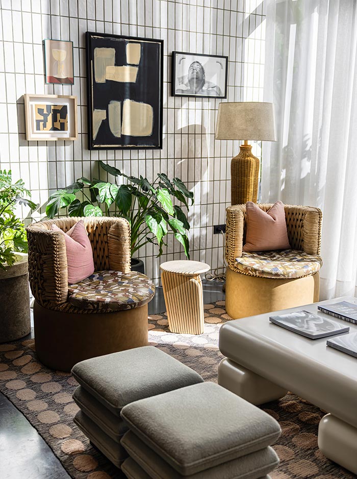 Cozy sitting area with natural light, featuring two woven armchairs with floral cushions and pink throw pillows, placed on a patterned carpet. A white modern coffee table and two cushioned stools sit in front, surrounded by leafy green plants and a golden wicker floor lamp. The tiled wall is decorated with abstract and black-and-white art in a casual gallery layout.