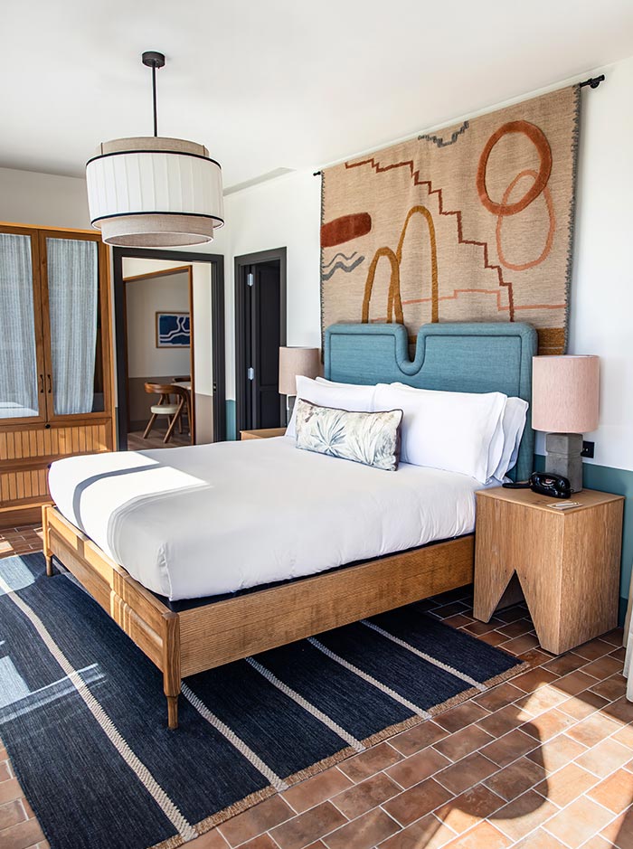 Bright, modern hotel room with a mid-century design, featuring a wooden bed frame with a white duvet and teal upholstered headboard. A large abstract wall hanging with geometric shapes in earth tones decorates the space above the bed, flanked by two wooden nightstands with pink-toned lamps. The room includes a navy striped rug on terracotta floor tiles and a large, round pendant light overhead.