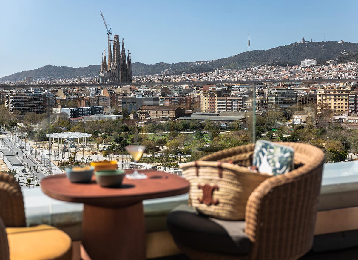Scenic rooftop view overlooking Barcelona with the iconic Sagrada Família basilica in the distance and hills in the background. In the foreground, a cozy outdoor seating area with wicker chairs, a small table with drinks and snack bowls, and a woven handbag resting on a chair. The glass railing provides an unobstructed view of the city and park below on a clear, sunny day.