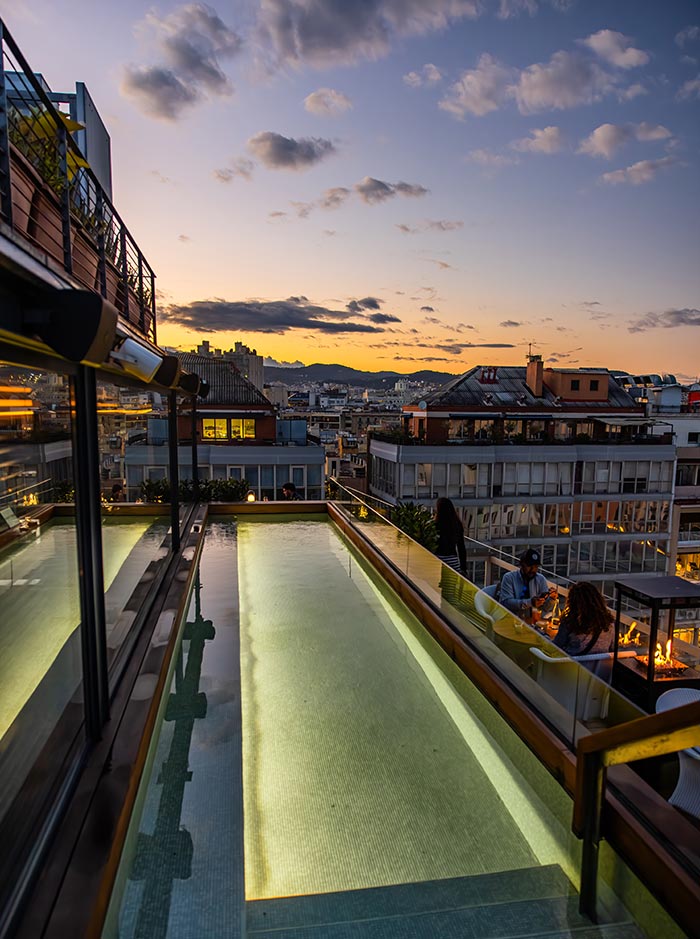 Rooftop pool at The Roof at Edition Barcelona, glowing softly with underwater lighting as the sun sets behind the city skyline. Surrounded by modern glass railings, the pool overlooks neighboring rooftops and distant hills under a colorful dusk sky. Guests relax nearby at a cozy firepit lounge area, enjoying the elevated ambiance.