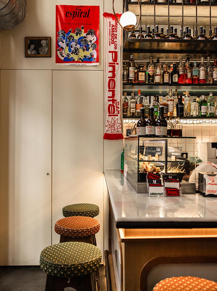 Interior of Bar Pimentel in Barcelona, showing a cozy bar setup with patterned cushioned stools, a marble counter, and shelves stocked with liquor bottles. A red scarf reading “Bar Pimentel” and a bold red poster titled “espiral” add pops of color to the warm, inviting atmosphere.