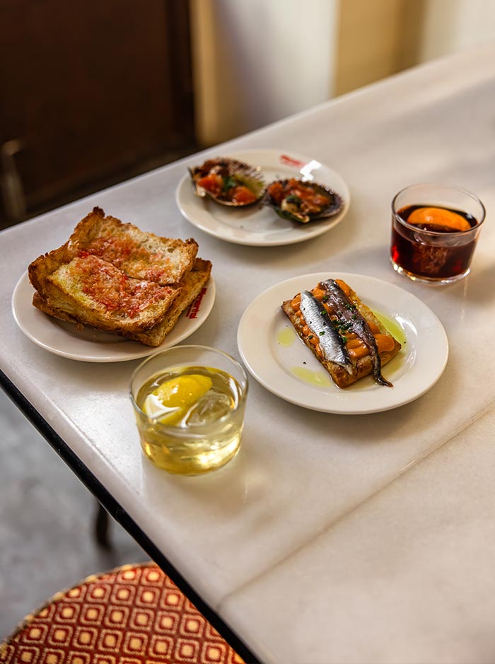 Tapas spread at Bar Pimentel in Barcelona, featuring pan con tomate, grilled shellfish with tomato, and anchovies on toast drizzled with olive oil, accompanied by a vermouth with orange and a cocktail with lemon over ice, all set on a marble table.