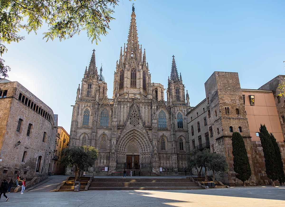 Front view of the Barcelona Cathedral (Cathedral of the Holy Cross and Saint Eulalia), showcasing its grand Gothic architecture with intricate stone carvings, pointed arches, and multiple spires. The surrounding plaza is quiet with a few people walking and historic stone buildings framing the scene under a clear blue sky.