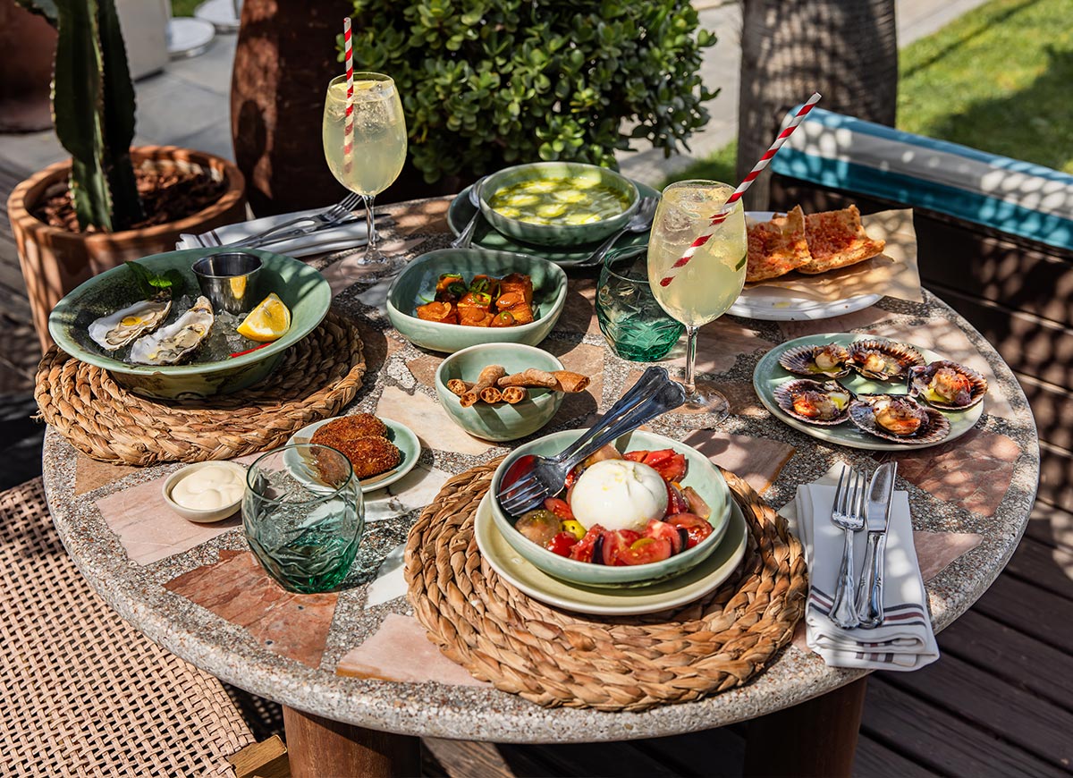 Colorful Mediterranean spread at Julieta’s restaurant in Barcelona, with dishes including oysters on ice, burrata with tomatoes, scallops in shell, crispy croquettes, pan con tomate, and patatas bravas. Two spritz cocktails with red striped straws sit among green ceramic tableware on a mosaic-topped outdoor table in the sun.