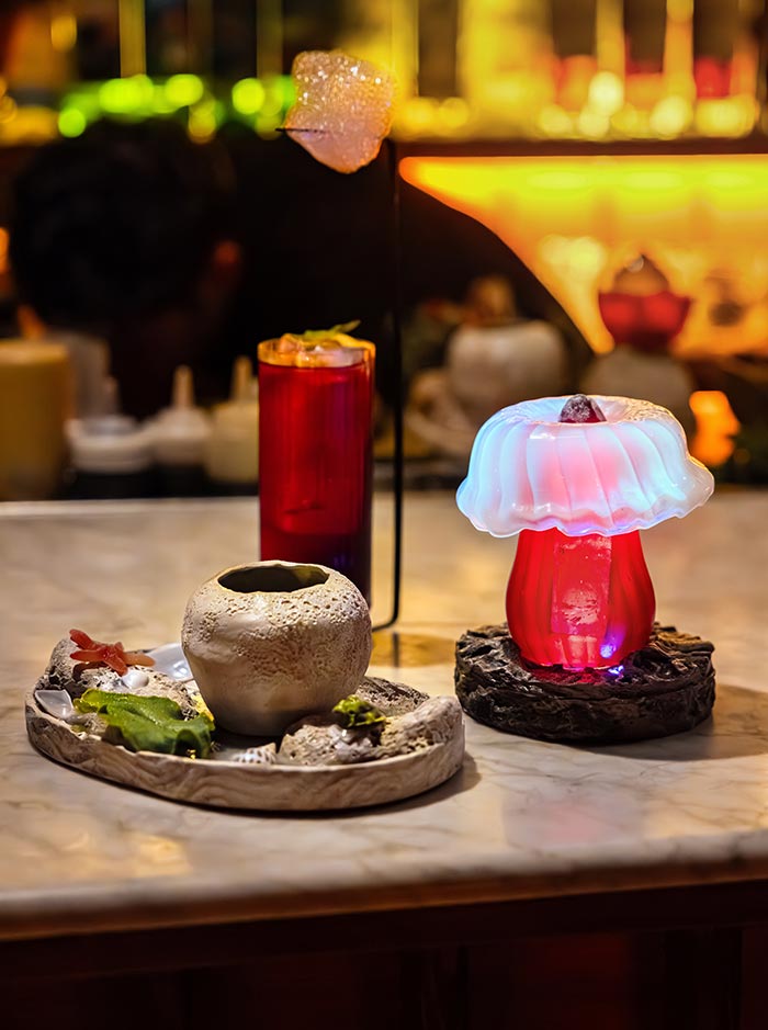 Close-up of three artistic cocktails at Paradiso in Barcelona, presented on a marble bar counter. One drink is served in a glowing red glass shaped like a mushroom with a frosted cap, another in a textured ceramic vessel on a tray with edible garnishes, and a tall red glass topped with a sugar bubble. The creative presentation highlights the bar's theatrical and experimental style.