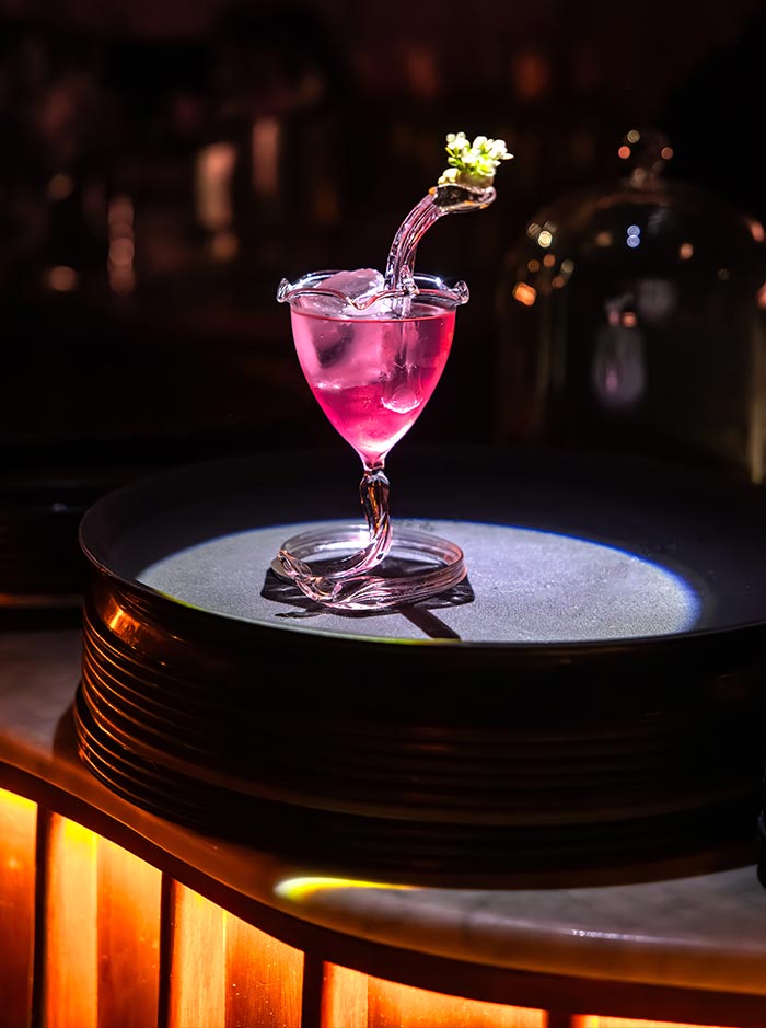 A spotlight highlights a sculptural pink cocktail in a twisted glass with a curved stem, served at Paradiso speakeasy bar in Barcelona. The drink features a translucent pink hue and is garnished with a delicate white flower on a small branch. The dramatic lighting and elegant presentation emphasize the bar’s flair for artistic mixology.