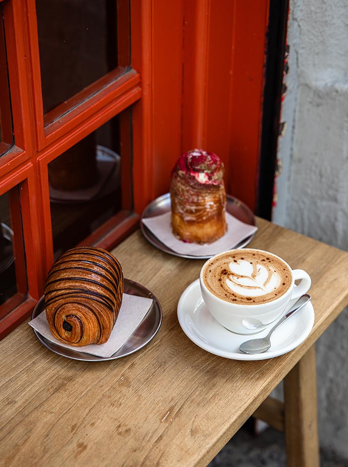 Close-up of a cozy café setup in Barcelona, featuring a beautifully layered pain au chocolat, a glazed pastry topped with pink cream, and a cappuccino with delicate latte art, all served on a wooden bench by a red-framed window.