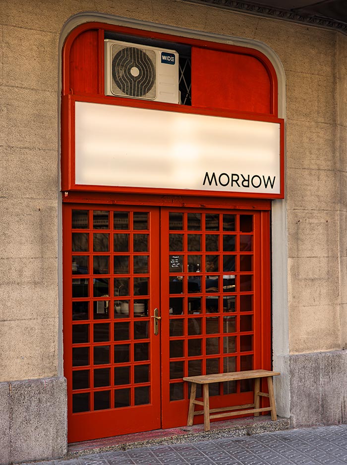 Street view of Morrow coffee shop in Barcelona, with bold red-framed glass doors and a small wooden bench out front. The sign above the entrance displays the word “MORROW” upside down, adding a quirky design touch to the minimalist facade.