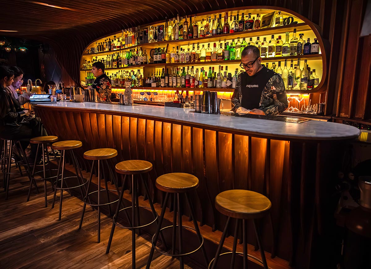 Dimly lit speakeasy-style bar with a sculpted wood bar front, glowing amber lights, and a marble countertop lined with wooden stools. Behind the bar, shelves are filled with an extensive selection of liquor bottles, framed by a curved wood structure. Bartenders in floral jackets and dark shirts prepare drinks while a couple of guests browse the menu.