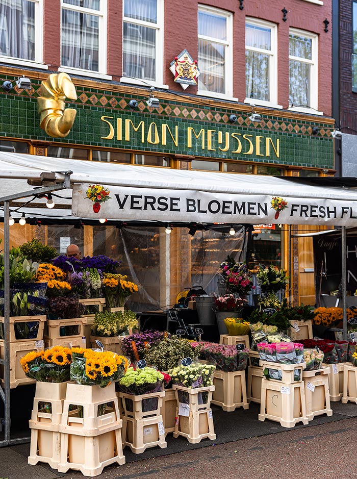 Flower stall at Albert Cuyp Market in Amsterdam with buckets of fresh blooms including sunflowers, roses, lilies, and mixed bouquets under a canopy reading “Verse Bloemen – Fresh Flowers.” The background shows the Art Deco-style green-tiled facade of Simon Meijssen bakery, adding local charm to the lively market scene.