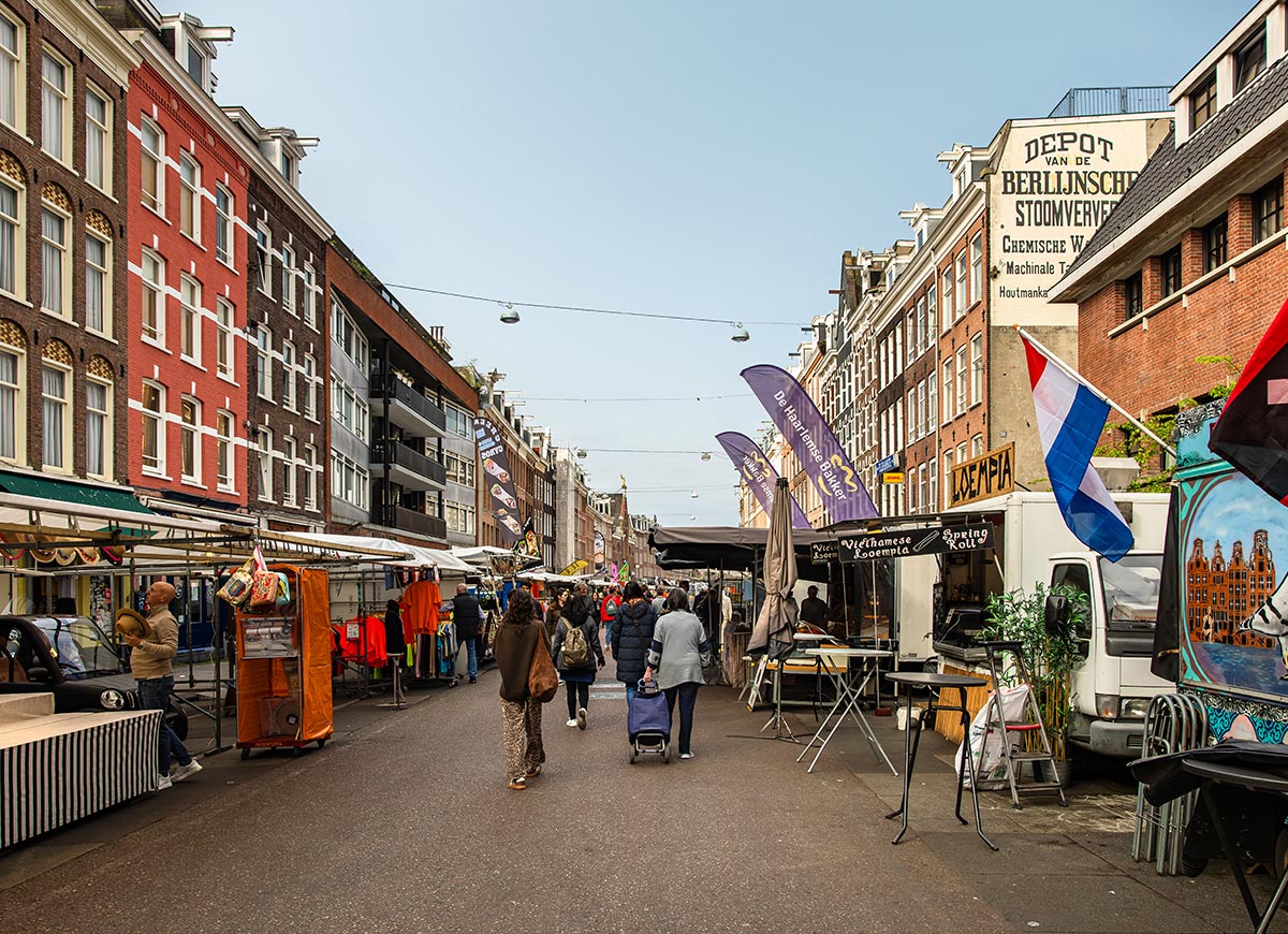 Bustling scene at Albert Cuyp Market in Amsterdam, with shoppers strolling between colorful street stalls selling clothing, produce, and snacks. Flags and vendor banners, including one for Vietnamese spring rolls, add cultural flair, while traditional Dutch architecture frames the lively open-air market. A clear sky and relaxed crowd reflect a typical day at one of the city's most iconic markets.







