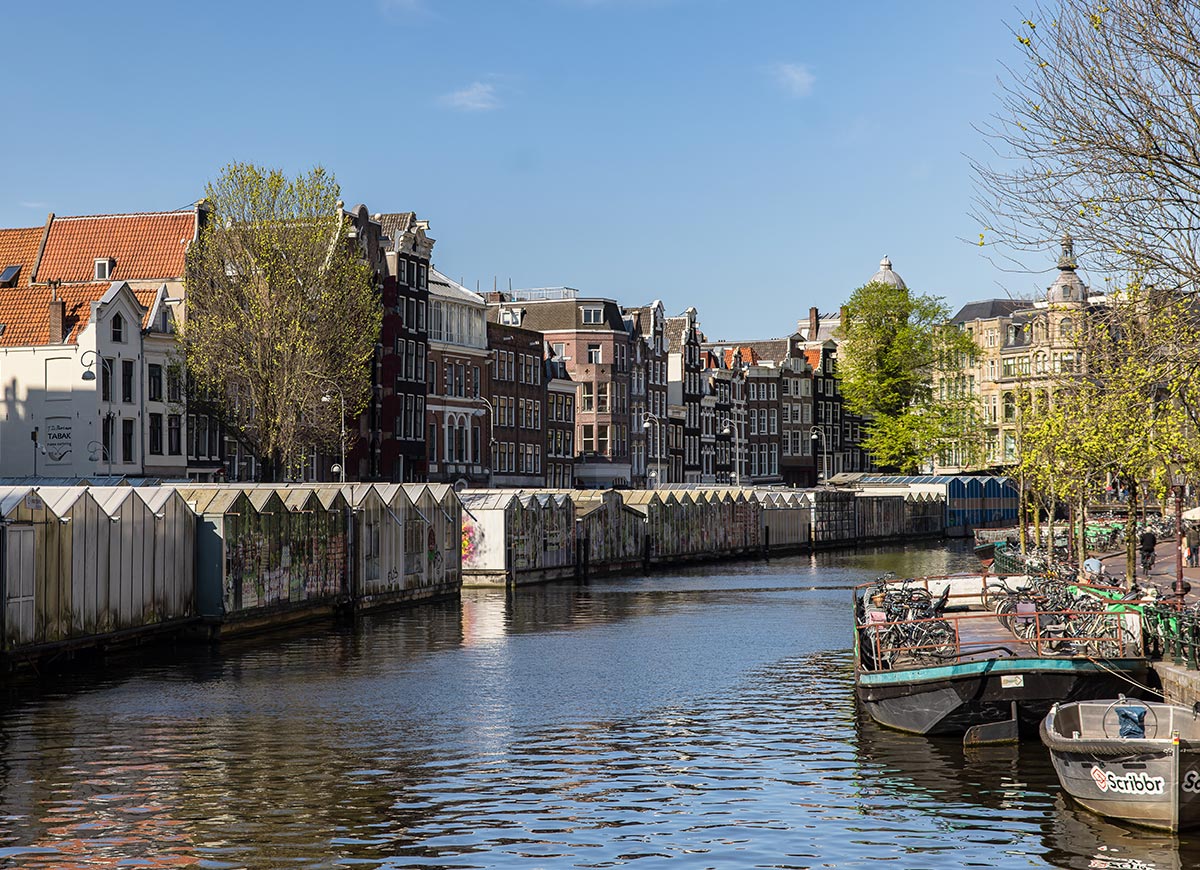 View of the Bloemenmarkt in Amsterdam, the world’s only floating flower market, with greenhouse-like stalls lining the canal. Traditional Dutch townhouses rise in the background under a clear blue sky, and boats with bicycles docked alongside the water add to the city's charm. Trees with fresh green leaves hint at springtime.







