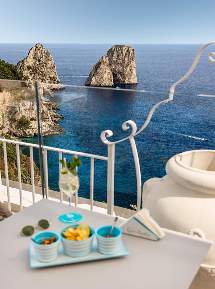 Table set with snacks and a cocktail at Capri Rooftop Lounge Bar, overlooking the stunning Faraglioni rock formations and sparkling blue sea from a high vantage point with white railings and coastal cliffs in the background.