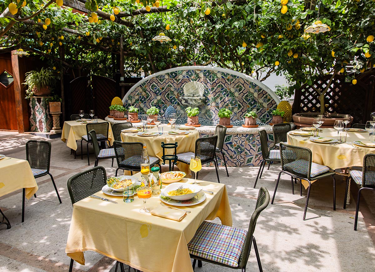 Charming outdoor dining area at Da Paolino restaurant in Capri, Italy, set beneath a canopy of lemon trees with tables dressed in yellow linens, colorful ceramic decor, and plates of fresh Italian cuisine.





