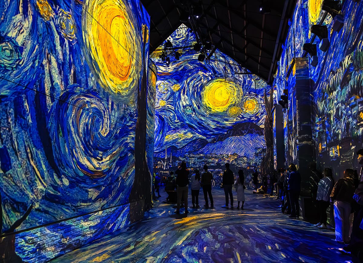 Immersive projection of Van Gogh’s "Starry Night" at Fabrique des Lumières in Amsterdam, enveloping the walls and floor in swirling blues and vivid yellows. Visitors are silhouetted against the luminous artwork, creating a dreamlike, immersive atmosphere. The digital display transforms the iconic painting into a full sensory experience.







