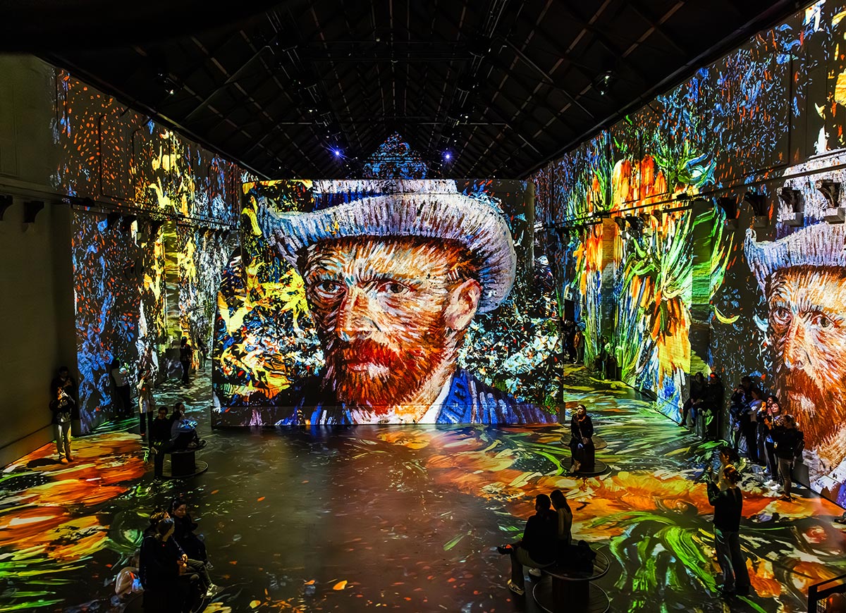 Immersive art exhibition at Fabrique des Lumières in Amsterdam, featuring a vivid digital projection of Vincent van Gogh’s self-portrait across towering walls and the floor. The space glows with swirling brushstrokes, floral motifs, and bold colors, enveloping visitors in a dynamic, 360-degree visual experience. Guests are seated and walking through the installation, capturing photos and taking in the animated artwork.


