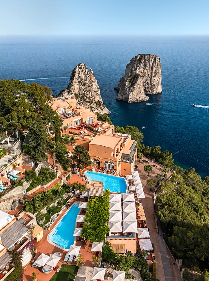 Aerial view of Hotel Punta Tragara in Capri, Italy, with its terracotta buildings, luxurious pools, and white sun umbrellas perched on a cliffside overlooking the iconic Faraglioni sea stacks and sparkling blue Mediterranean.