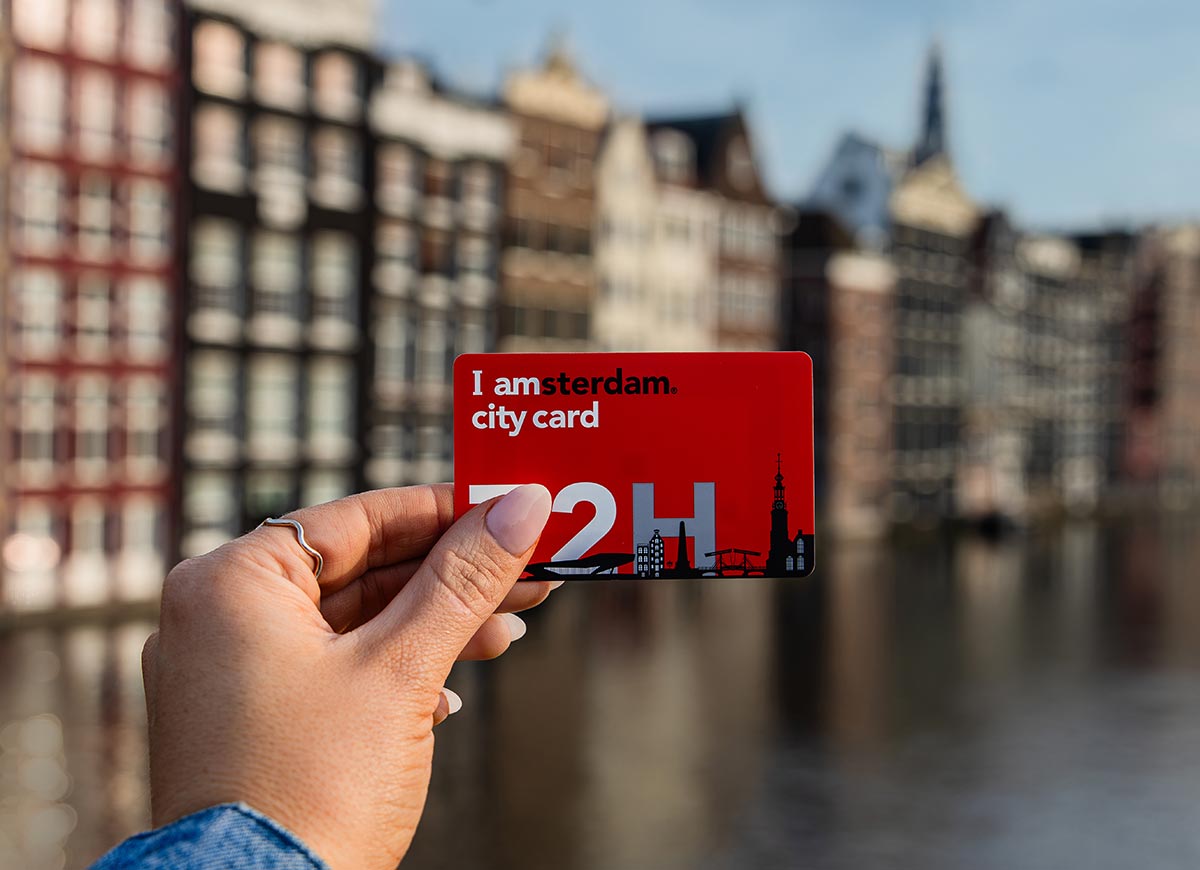 A close-up of a hand holding the red "I amsterdam City Card" marked with "72H" in bold white letters, with the iconic canal houses of Amsterdam softly blurred in the background. The card includes black silhouettes of Amsterdam landmarks.







