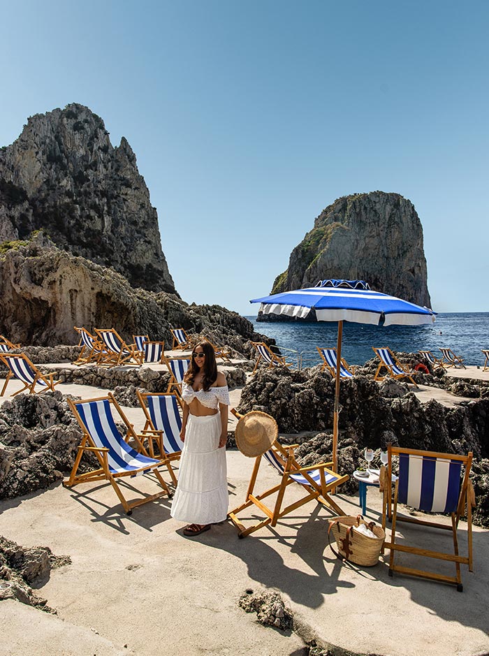Woman in a white outfit standing among blue-and-white striped beach chairs and umbrellas at La Fontelina Beach Club in Capri, Italy, with dramatic rock formations and the Faraglioni in the background.