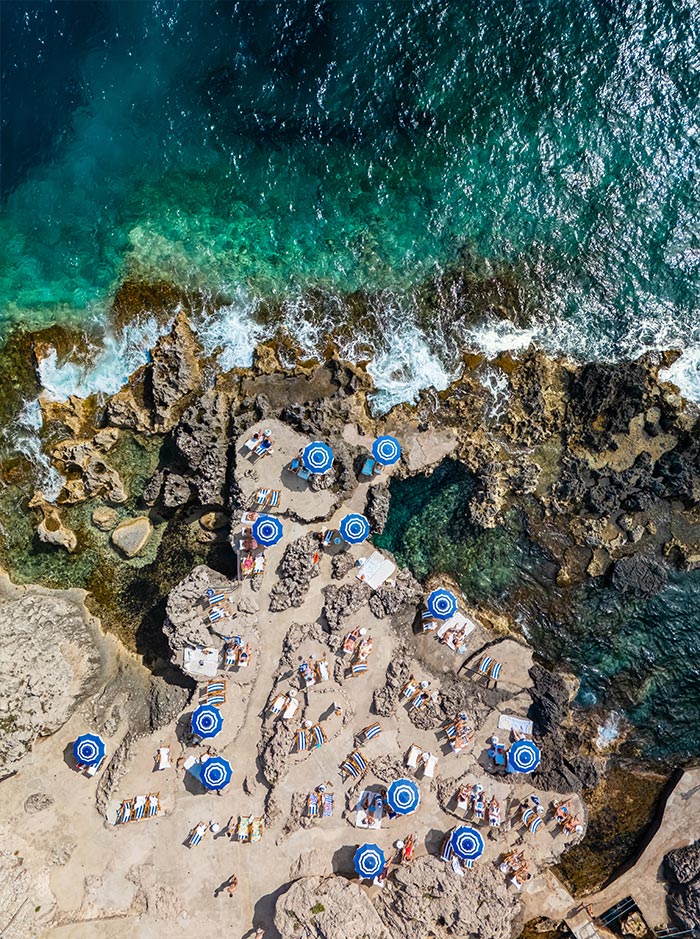 Aerial view of La Fontelina Beach Club in Capri, Italy, with blue-and-white striped umbrellas and sunbeds arranged across rugged rock formations beside the vibrant turquoise and deep blue sea.