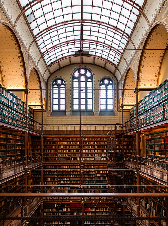 Interior of the Cuypers Library at the Rijksmuseum in Amsterdam, featuring tall shelves filled with books that rise up multiple stories beneath an arched glass ceiling. The historic space is lined with ornate iron railings and spiral staircases, with natural light filtering through large stained-glass windows. It exudes a timeless, scholarly atmosphere.