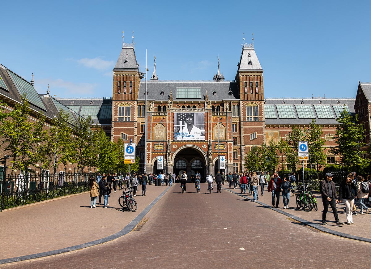 Front view of the Rijksmuseum in Amsterdam, showcasing its grand Neo-Gothic architecture with twin towers and arched central passage. A large banner for an exhibition hangs above the entrance as visitors walk and cycle along the tree-lined path leading up to the museum. The clear sky and lively crowd highlight the popular cultural destination.

