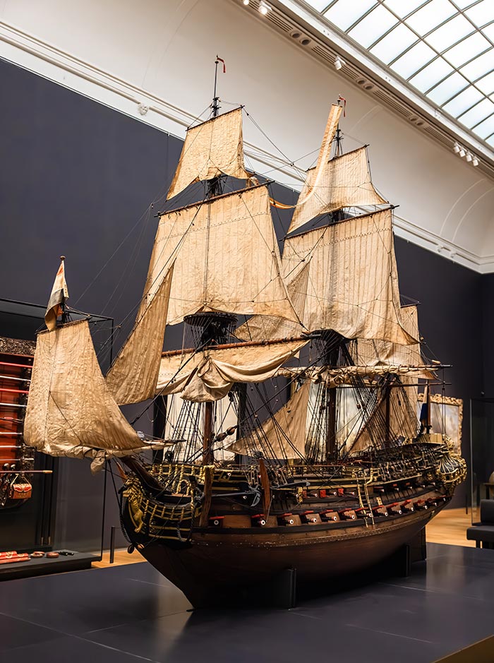 Large, detailed model of an 18th-century Dutch warship displayed at the Rijksmuseum in Amsterdam. The ship features multiple sails, intricate rigging, and ornate gold accents, with rows of cannons visible along the hull. Set in a dark-walled gallery with overhead lighting, the exhibit highlights Dutch maritime history.