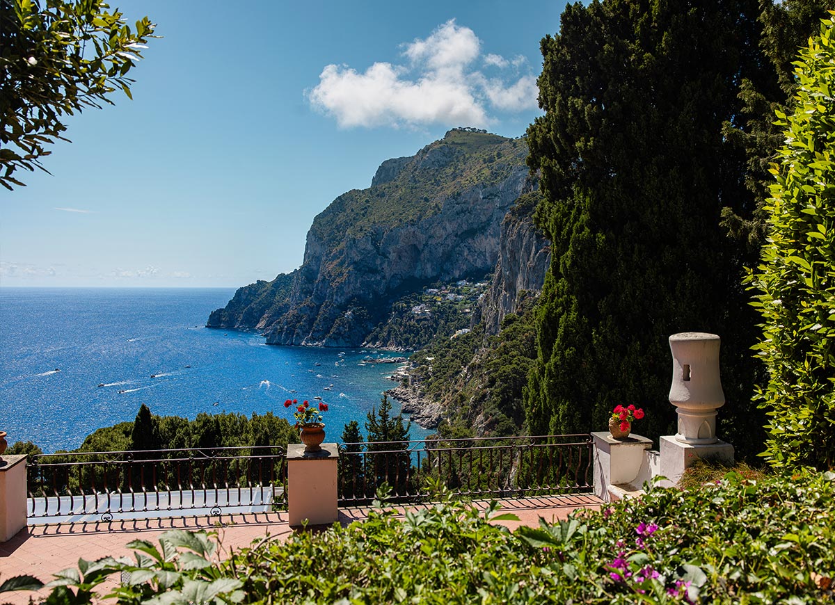 Scenic coastal view of Capri, Italy, with a terrace garden overlooking the vibrant blue sea, boats in the water, and dramatic cliffs rising in the background under a sunny sky.