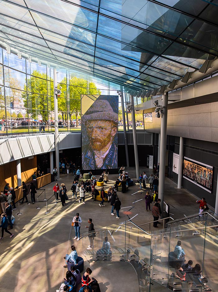 Interior of the Van Gogh Museum in Amsterdam, showing a bustling atrium with visitors walking, sitting, and queuing beneath a massive portrait of Vincent van Gogh. The space features a high glass ceiling, letting in natural light and offering views of the green park outside. A modern staircase and exhibit signage add to the museum's contemporary design.