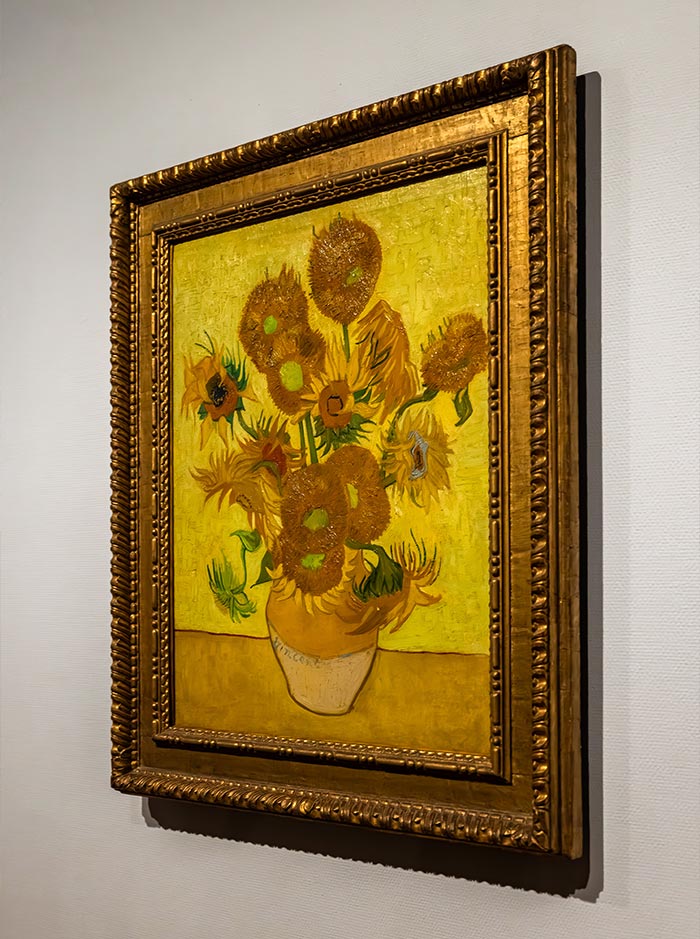 Framed painting of Vincent van Gogh’s famous “Sunflowers” displayed at the Van Gogh Museum in Amsterdam. The artwork features a vibrant yellow background with a vase of golden sunflowers in various stages of bloom and decay, rendered in Van Gogh’s signature expressive brushstrokes. The ornate gold frame enhances the bold, textured composition.