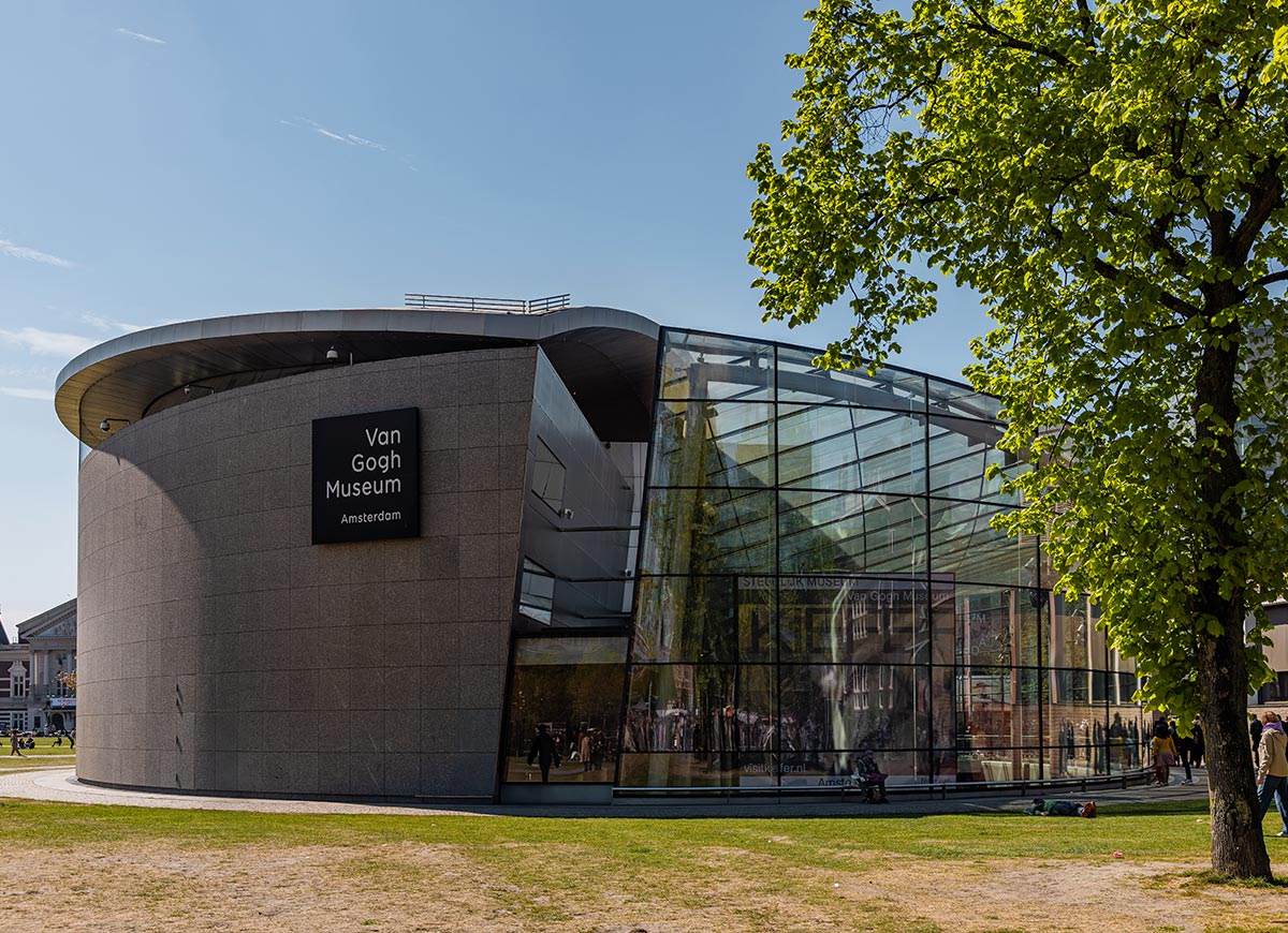 Exterior of the Van Gogh Museum in Amsterdam, featuring a sleek modern design with a curved stone façade and a large glass-walled atrium. The museum sign is prominently displayed, and visitors are seen gathering near the entrance under clear blue skies. A leafy tree and grassy lawn add a touch of greenery to the scene.







