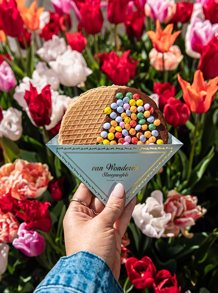 Hand holding a Van Wonderen Stroopwafel topped with colorful candy-coated chocolates, partially wrapped in branded blue paper. The background is filled with vibrant blooming tulips in shades of red, pink, orange, and white, capturing a festive springtime moment in Amsterdam.