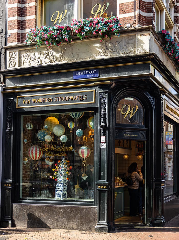 Charming storefront of Van Wonderen Stroopwafels on Kalverstraat in Amsterdam, with a black and gold exterior and large display windows featuring whimsical hot air balloon decor. A colorful flower arrangement sits in the window alongside a tall blue-and-white ceramic vase. Customers are seen inside the cozy shop, known for its artisanal Dutch stroopwafels.