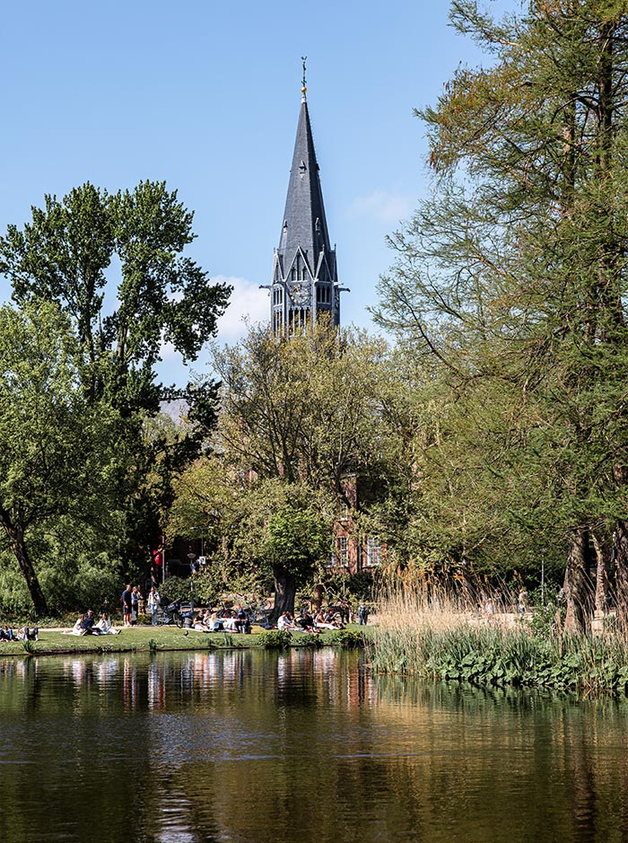 Serene view of Vondelpark in Amsterdam with people relaxing along the grassy edge of a pond, surrounded by trees and tall grasses. In the background, the pointed spire of the Vondelkerk church rises above the treetops under a clear blue sky. The peaceful setting reflects in the still water, adding to the park's tranquil charm.