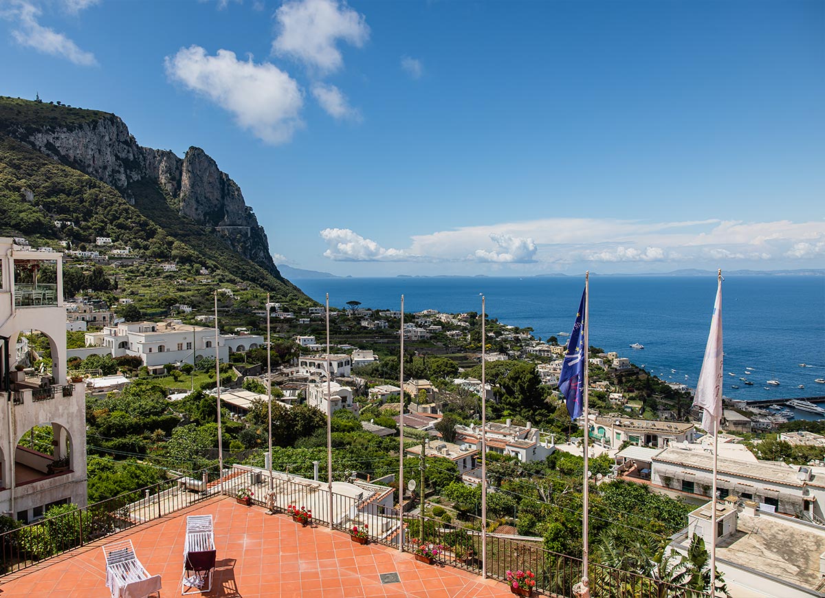 Panoramic view of Capri Island, Italy, from a terracotta rooftop terrace with lounge chairs and flagpoles, overlooking whitewashed buildings, green hillsides, and the sparkling blue sea stretching to the horizon.






