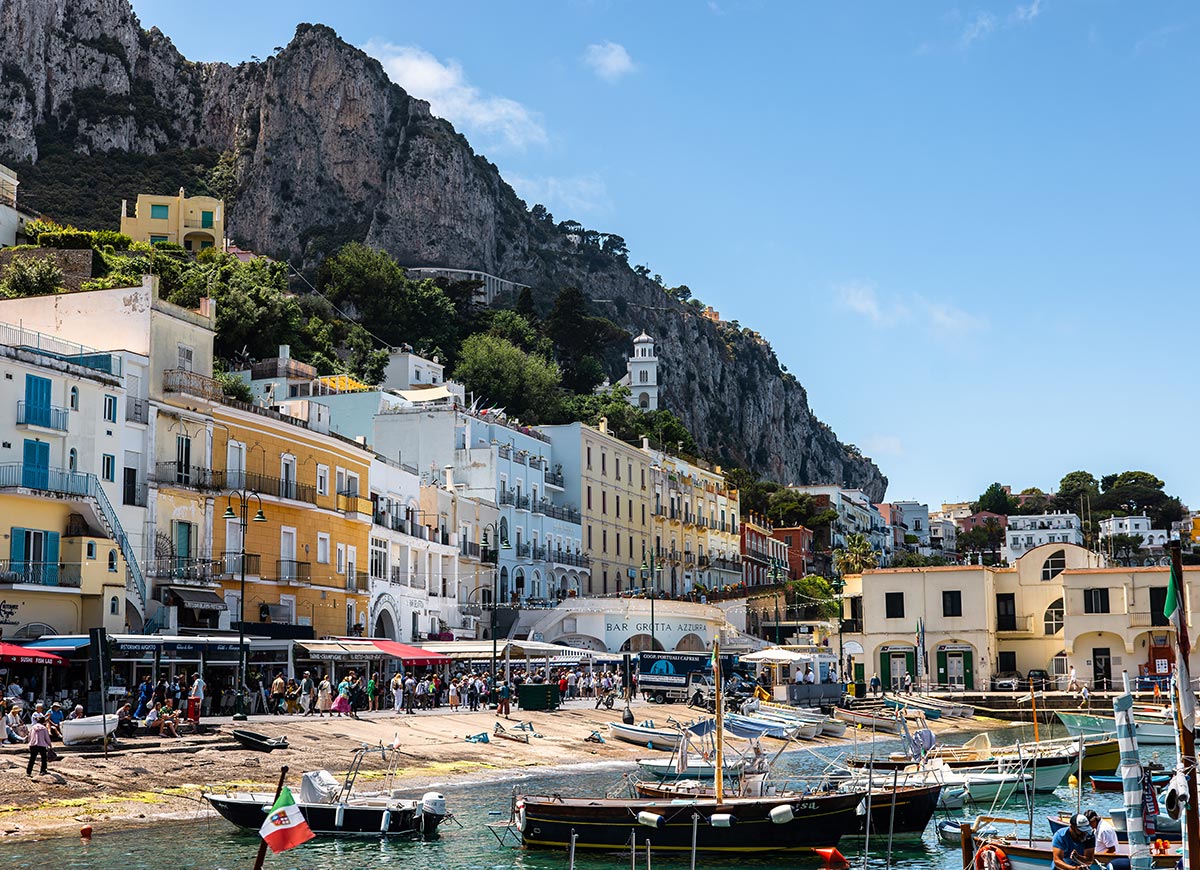 Bustling scene at the main port of Capri, Italy, with colorful buildings stacked along the hillside, small boats docked in the turquoise water, and crowds gathered along the waterfront beneath a dramatic rocky cliff.






