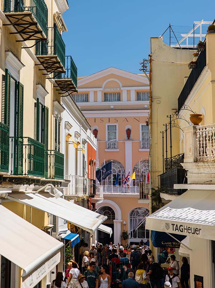 Bustling pedestrian street in Capri town, Italy, lined with colorful buildings, designer shops like TAG Heuer, and sunshades, leading toward the peach-toned Grand Hotel Quisisana with flags flying from its terrace.