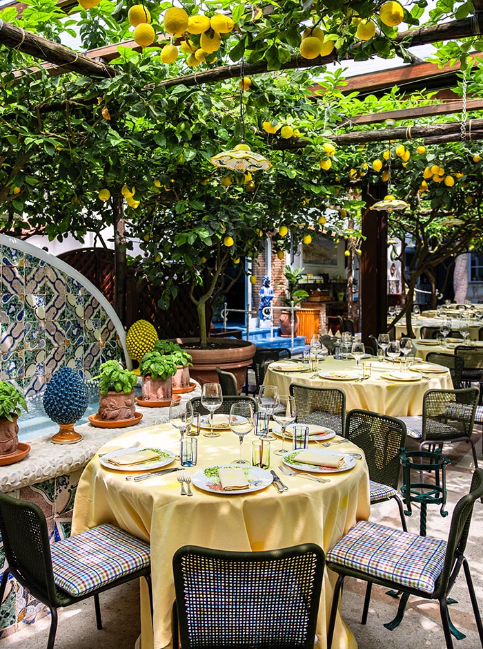 Lush outdoor dining area at Da Paolino restaurant in Capri, Italy, with yellow-clothed round tables set beneath lemon trees, surrounded by colorful tile decor, potted basil, and hanging ceramic lamps.