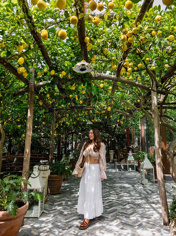 Woman walking under a pergola of lush lemon trees at the entrance of Da Paolino restaurant in Capri, Italy, surrounded by potted plants, rustic lanterns, and dappled sunlight.