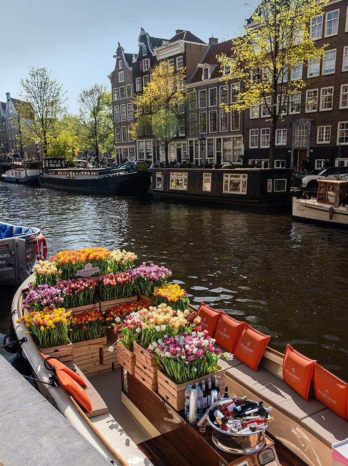 Tulip-decorated Flagship Amsterdam boat docked on a canal, filled with vibrant wooden crates of fresh tulips in shades of pink, yellow, orange, and white. Orange branded cushions line the seats, and a drink station with bottles and an ice bucket adds a festive touch. Traditional Dutch canal houses and houseboats create a charming backdrop under a sunny sky.