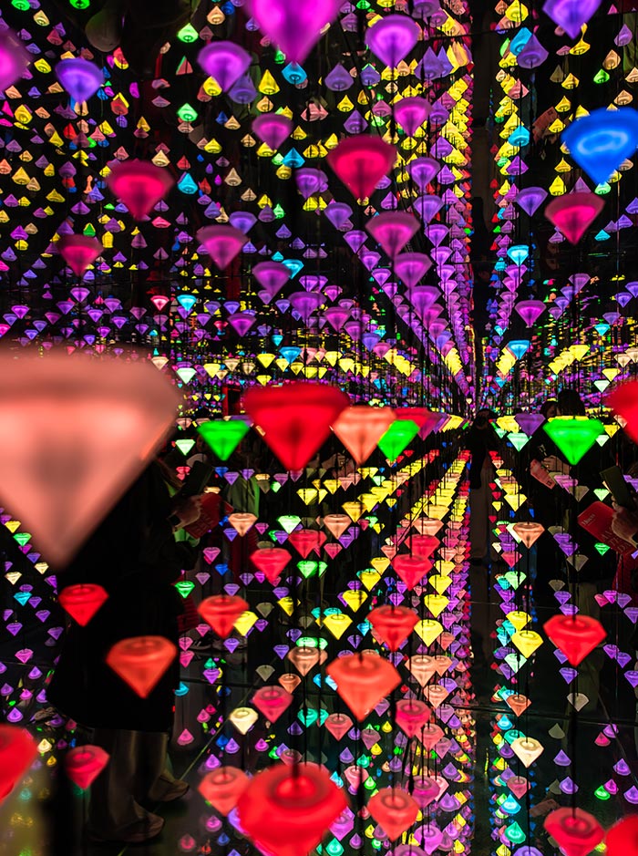 Vibrant installation at the Moco Museum in Amsterdam featuring hundreds of suspended, glowing diamond-shaped lights in a rainbow of colors. The mirrored walls and floor create an infinite reflection effect, immersing visitors in a dazzling, kaleidoscopic environment. Silhouettes of guests can be seen interacting with the mesmerizing exhibit.