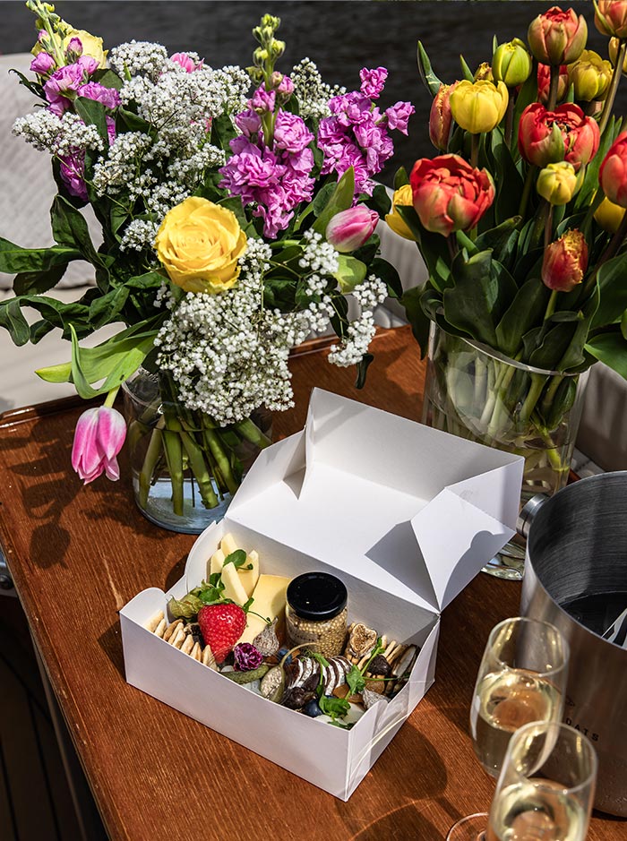 Close-up of a gourmet snack box on a wooden boat table in Amsterdam, filled with cheese, crackers, a strawberry, fig slices, and a small jar of mustard. The table is also decorated with vibrant floral bouquets of tulips, roses, and baby's breath, alongside champagne glasses and an ice bucket. A charming and elegant canal cruise setup.