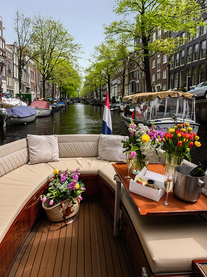 Cozy open-air boat cruising through an Amsterdam canal, with tan cushioned seating, floral bouquets, champagne, and snacks set on a wooden table. The Dutch flag waves at the boat’s stern as leafy green trees and historic canal houses line the waterway under a clear sky. A wicker bag filled with fresh flowers adds a charming touch to the elegant setup.