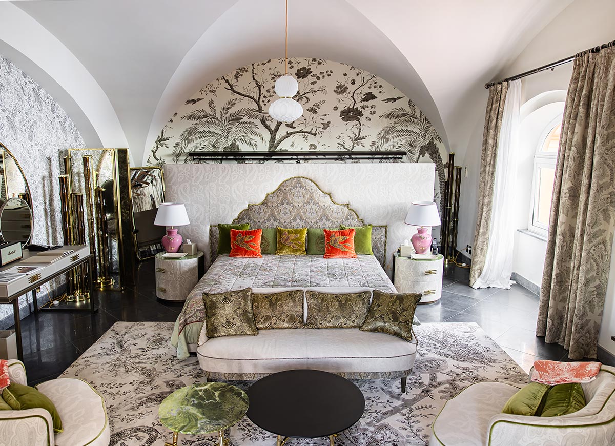 Elegant suite at Hotel Punta Tragara in Capri, Italy, featuring a vaulted ceiling, intricate botanical wallpaper, a lavish king-sized bed with colorful accent pillows, and a mix of vintage and modern furnishings in soft, neutral tones.